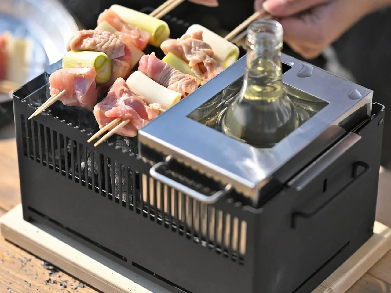 Skewers of meat and green onion on a small black tabletop grill, with a bottle warming in a stainless insert beside them