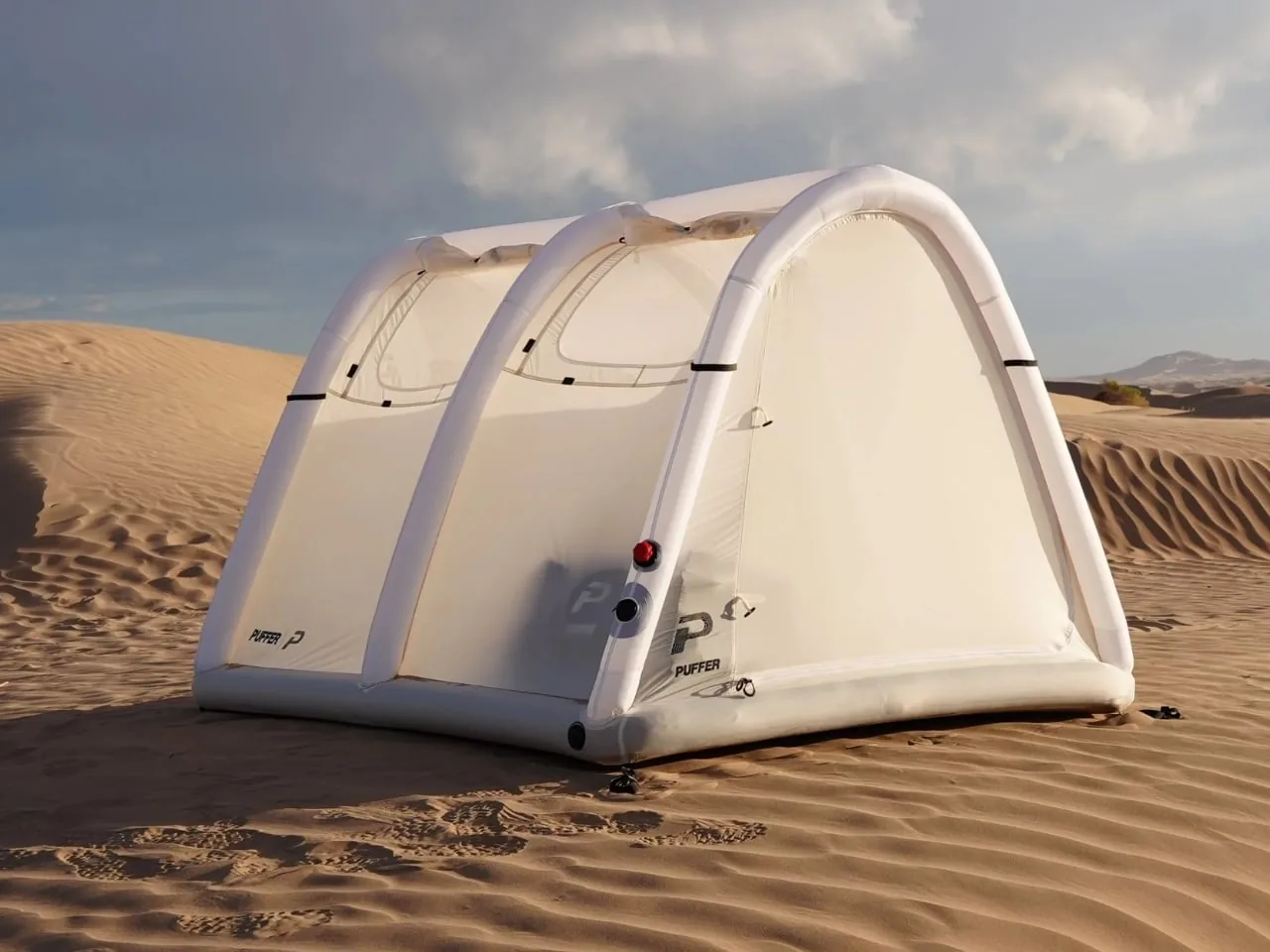 A white inflatable Puffer tent pitched on sand dunes under a cloudy sky