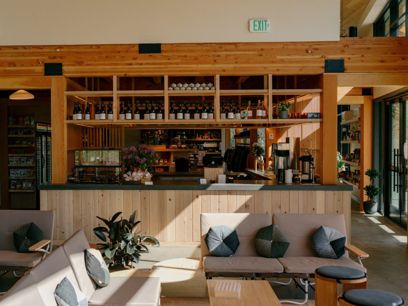 Inside the Campstore, a pine cafe counter under open shelves of wine bottles, with low camp sofas in the sunlit foreground