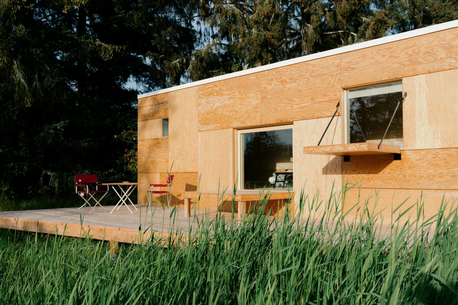 A Jyubako Suite at Snow Peak Campfield, a small pale plywood cabin with a wide window and a fold-down ledge, on a deck with two red camp chairs in tall grass