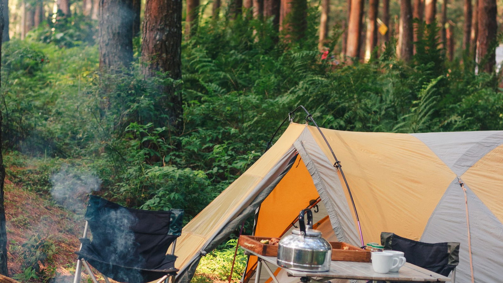 A tent pitched in a green forest with a kettle, camp chairs and a cook table set up beside it