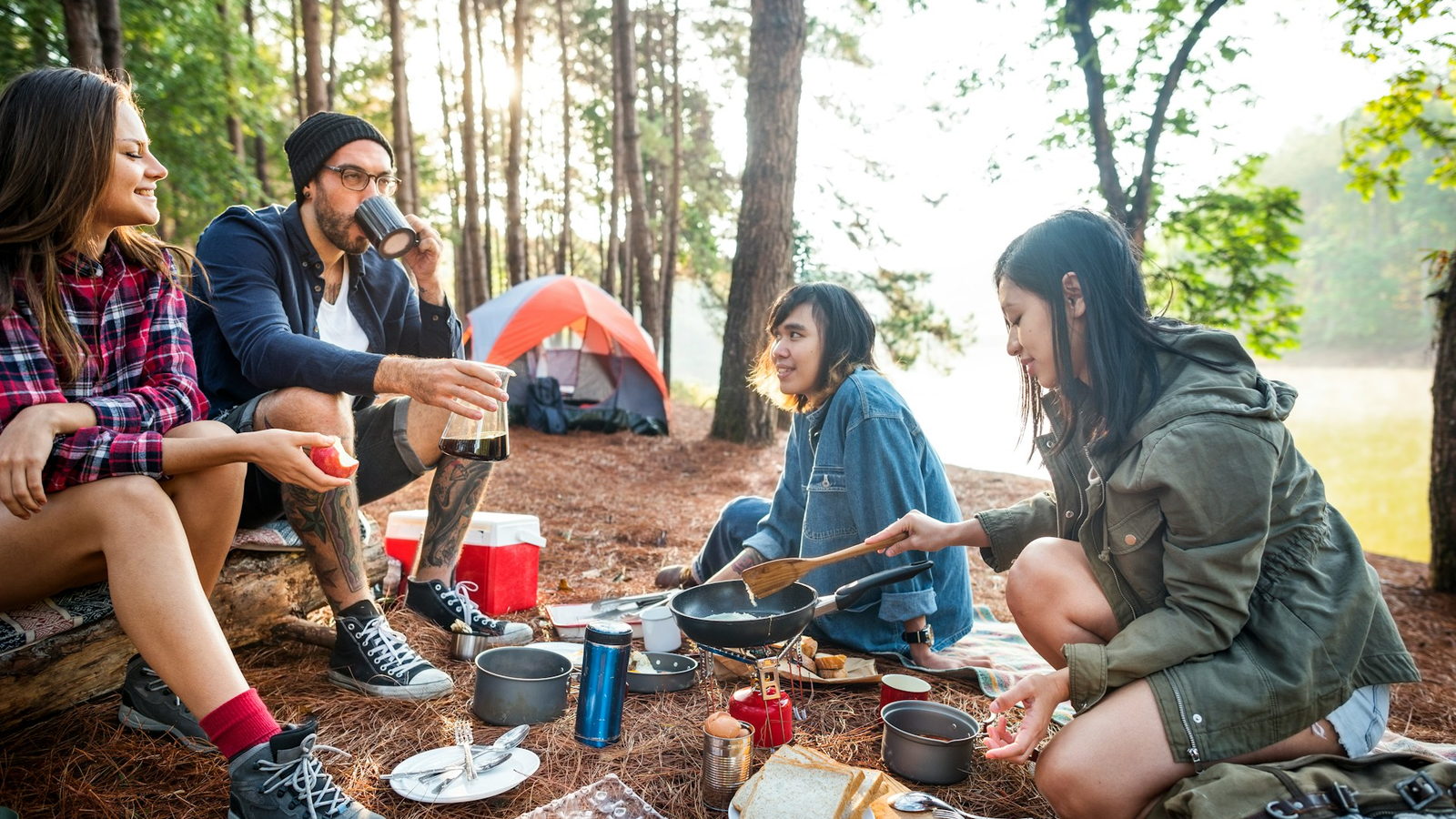 Four friends cooking breakfast at a campsite under pines, a tent behind them