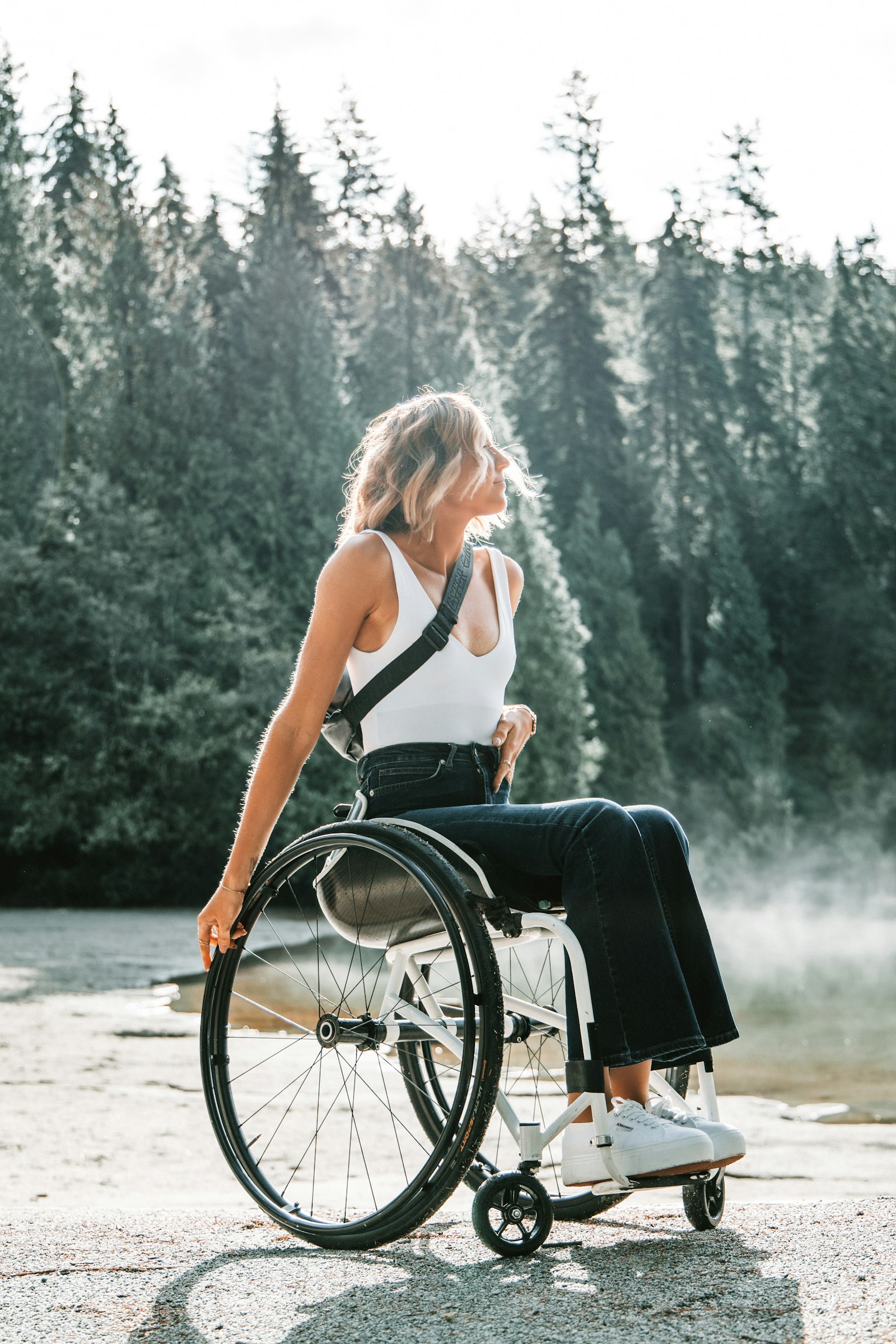 A woman in a wheelchair pauses at the edge of a misty lake at dawn, surrounded by tall pines